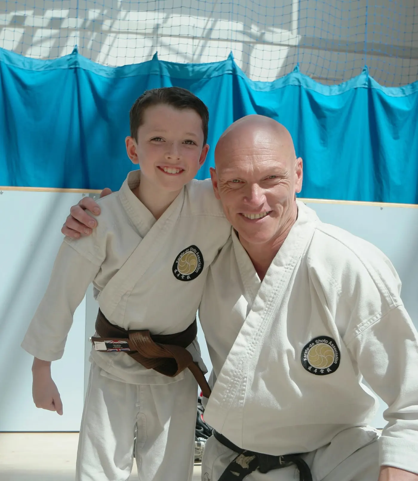 A smiling junior with a new brown belt, his instructor's arm around his shoulder