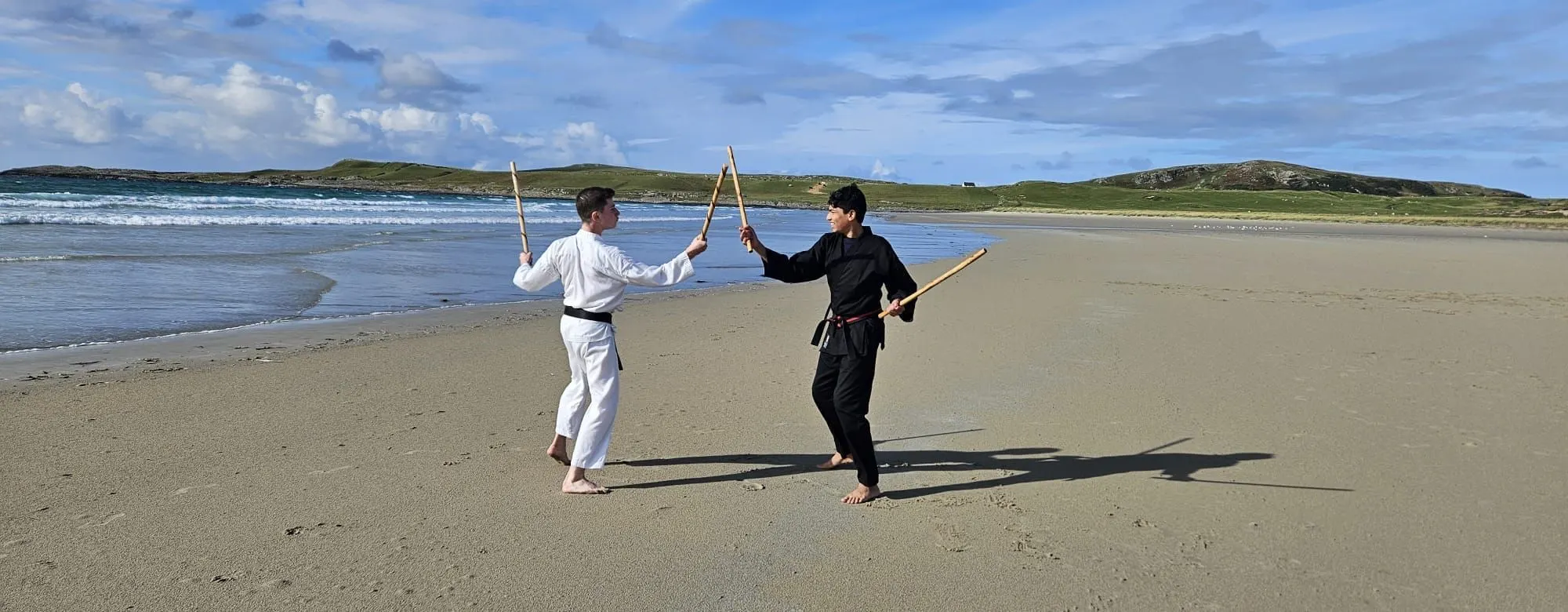 Two members practising with bo staffs on a beach